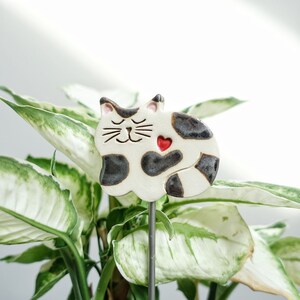 Könnte beinhalten: Eine Keramik-Katzenfigur mit schwarzen und weißen Flecken und einem roten Herzen auf der Brust. Die Katze ist in einer schlafenden Position zusammengerollt. Die Figur ist an einem Metallpfahl befestigt, wodurch sie sich gut als Gartendekoration eignet.