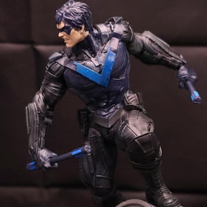 May include: A black and blue action figure of a superhero standing on a gray base with a bat symbol. The figure is holding a blue weapon in its right hand.