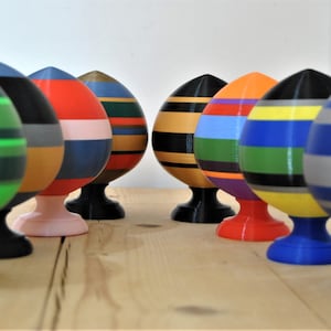 Puede incluir: Una colección de diez adornos coloridos en forma de huevo, rayados, con una parte superior redondeada y una base. Los adornos están hechos de un material liso y duro y tienen una variedad de colores, incluyendo verde, rojo, azul, amarillo, negro y blanco.