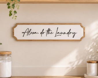May include: A rectangular wall sign with a light wood frame and a white background featuring the black cursive text Alexa, do the Laundry. The sign is mounted on a wall above a wooden countertop with laundry appliances and various home decor items.