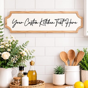 May include: A rectangular wall sign with a light wood frame and a white centre panel featuring the black script text Your Custom Kitchen Text Here. The sign has decorative curved edges on the left and right sides and is mounted on a white tiled wall.