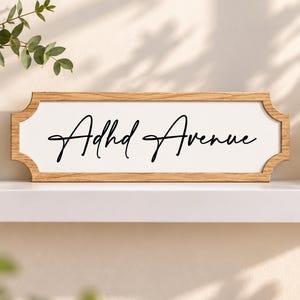 May include: A rectangular wooden sign with a light oak finish and a white centre panel. The sign features the black cursive text Adhd Avenue centred on the white background. The frame has decorative notched corners on the left and right sides.