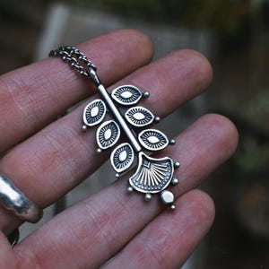 May include: Silver-toned pendant necklace featuring a leaf-like design. The pendant has a central stem with oval-shaped elements and a fan-shaped base. The necklace is displayed on a hand, showcasing the intricate details of the jewelry.