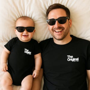 Może przedstawiać: Ojciec i syn w dopasowanych czarnych koszulkach z białym nadrukiem. Koszulka ojca ma napis "The Original EST 1984", a koszulka syna "The Remix EST 2024". Oboje noszą czarne okulary przeciwsłoneczne.