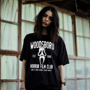 Op de afbeelding: Zwart T-shirt met een witte afbeelding van een spookgezicht en de tekst "Woodsboro Est. 1996 Horror Film Club Wat is je favoriete horrorfilm?"