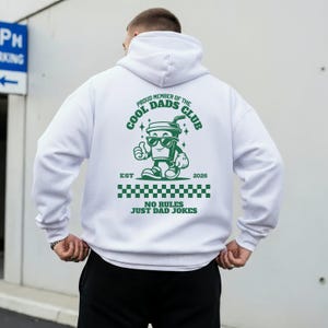 Könnte beinhalten: Ein weißer Kapuzenpullover mit einer grünen Grafik auf der Rückseite. Das Design zeigt einen Cartoon-Becher mit Sonnenbrille, den Text PROUD MEMBER OF THE COOL DADS CLUB, EST 2026, ein Schachbrettmuster und den Text NO RULES JUST DAD JOKES.