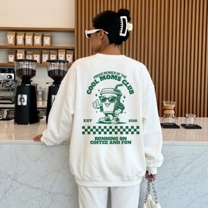 Könnte beinhalten: Cremefarbenes Sweatshirt mit einem grünen Aufdruck auf dem Rücken. Der Aufdruck zeigt den Text "PROUD MEMBER OF THE COOL MOMS CLUB" und eine Kaffeetassen-Figur. Der Text "RUNNING ON COFFEE AND FUN" ist ebenfalls aufgedruckt.