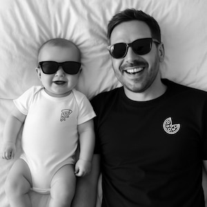 Op de afbeelding: Zwart-wit foto van een baby en een volwassene met een zonnebril. De baby draagt een witte romper met een pizzapunt-afbeelding. De volwassene draagt een zwart T-shirt met een pizzapunt-afbeelding.