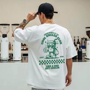 Könnte beinhalten: Weißes T-Shirt mit einem grünen Aufdruck einer Kaffeetasse mit Sonnenbrille und einem Daumen hoch. Der Text lautet "PROUD MEMBER OF THE COOL UNCLES CLUB" und "LIKE A DAD BUT COOLER". Das Design hat ein grünes Schachbrettmuster.