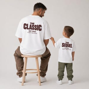 Op de afbeelding: Twee witte t-shirts met donkerbruine tekst op de achterkant. Het grotere shirt toont THE CLASSIC EST 1984 FIRST RELEASE, terwijl het kleinere shirt THE SEQUEL EST 2026 NEXT RELEASE toont. Beide shirts worden gedragen met een joggingbroek.