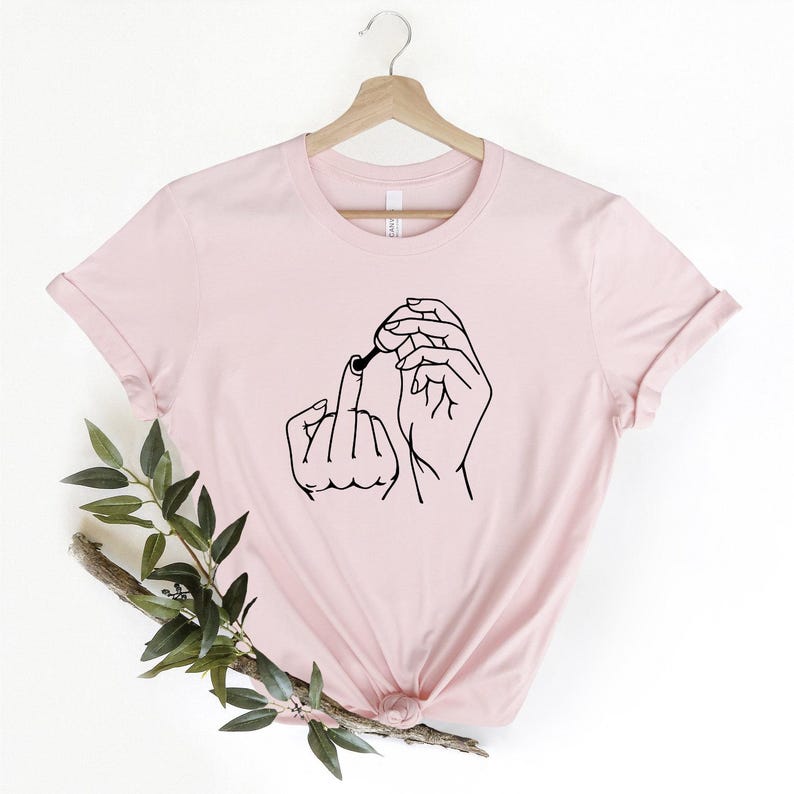 May include: A pink t-shirt with a black line drawing of two hands, one giving the middle finger and the other holding a nail polish bottle.