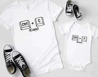 Pode incluir: Duas camisetas brancas com gráficos de teclas de teclado de computador pretas. A camiseta maior diz "Ctrl + C (Copiar)" e a camiseta menor diz "Ctrl + V (Colar)".