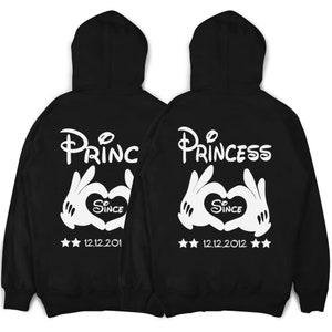 Könnte beinhalten: Zwei schwarze Kapuzenpullover mit weißem Text und Grafiken. Der linke Pullover sagt "Prince" mit einem Herzen und "Since 12.12.2011". Der rechte Pullover sagt "Princess" mit einem Herzen und "Since 12.12.2012".