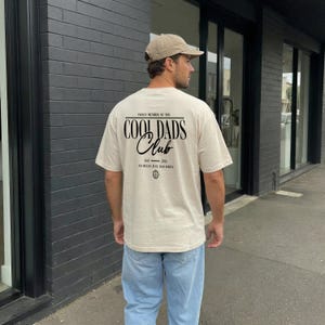 Op de afbeelding: Een beige t-shirt met korte mouwen en zwarte tekst op de achterkant met de tekst PROUD MEMBER OF THE COOL DADS CLUB EST 2026 NO RULES JUST DAD JOKES en een klein wereldbolicoon. Het shirt wordt gedragen met een lichtblauwe spijkerbroek en een beige pet.