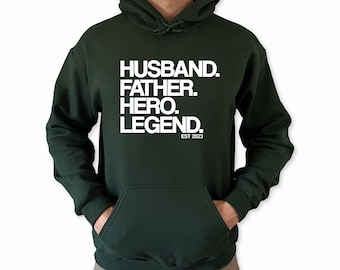 May include: Dark green hooded sweatshirt with white text that reads "Husband. Father. Hero. Legend. Est 2023."