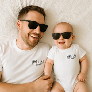 Könnte beinhalten: Ein lächelnder Erwachsener und ein Baby tragen schwarze Sonnenbrillen. Der Erwachsene trägt ein weißes T-Shirt mit einem "Ctrl + C (Kopieren)"-Aufdruck. Das Baby trägt einen weißen Body mit einem "Ctrl + V (Einfügen)"-Aufdruck.