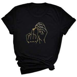 May include: Black t-shirt with a gold outline of a hand giving the middle finger and applying nail polish.