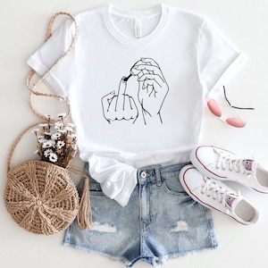 May include: White t-shirt with a black line drawing of two hands making the middle finger gesture. The shirt is worn with denim shorts and white sneakers.
