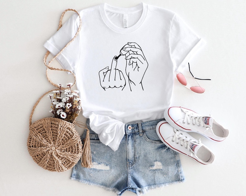 May include: White t-shirt with a black line drawing of two hands making the middle finger gesture. The shirt is worn with denim shorts and white sneakers.