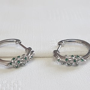 May include: A pair of silver hoop earrings with a twisted design and green gemstones.