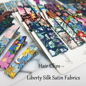 May include: A selection of patterned hair clips made from Liberty Silk Satin Fabrics. The clips display floral and other designs in shades of blue, pink, yellow, and green. The image includes the text "Hair Clips ~ Liberty Silk Satin Fabrics."