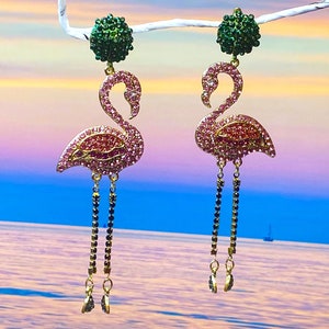 May include: Flamingo-shaped earrings with pink and red rhinestones. Each earring has a green rhinestone ball at the top and dangling chains with black and gold accents. The background shows a sunset over the sea. The earrings are a statement piece.