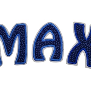 Könnte beinhalten: Blaue Filzbuchstaben, die den Namen "MAX" buchstabieren, mit einem weißen Umriss. Die Buchstaben sind gepolstert und haben eine gepunktete Textur.