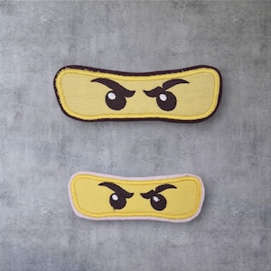 Könnte beinhalten: Zwei gestickte gelbe Ninja-Gesichtspatches mit schwarzen Augenbrauen und Augen. Die Patches haben eine rechteckige Form mit einem braunen und rosa Rand. Die Patches sind auf einem grauen Hintergrund platziert.
