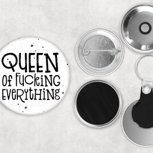 Könnte beinhalten: Ein weißer Button mit dem Text "QUEEN of FUCKING EVERYTHING" in Schwarz, mit schwarzen Tintenspritzern. Enthält eine Anstecknadel, eine Magnetrückseite und einen Schlüsselanhänger.