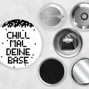 Könnte beinhalten: Ein weißer Knopf mit schwarzem, pixelartigem Text, der "CHILL MAL DEINE BASE" lautet. Der Knopf ist von sechs weiteren Knöpfen umgeben, darunter ein leerer schwarzer Knopf, ein silberner Knopf mit einem Flaschenöffner und ein silberner Knopf mit einer Stecknadel.