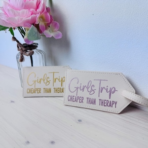 Könnte beinhalten: Zwei cremefarbene Gepäckanhänger mit dem Text "Girls Trip CHEAPER THAN THERAPY" in goldener und violetter Schrift. Die Anhänger haben eine strukturierte Oberfläche und einen Riemen. Im Hintergrund eine Vase mit rosa Blumen.