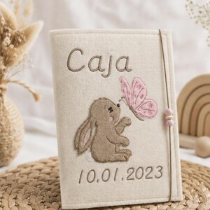 Funda de fieltro para el carné de salud infantil o de maternidad, bordada con un conejito y una mariposa. Personalizable con el nombre y, si se desea, la fecha de nacimiento.