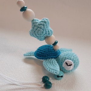 Puede incluir: Un juguete de tortuga de crochet azul y blanco con una estrella y cuentas de madera colgando de una cuerda blanca. La tortuga tiene una campana unida a su caparaz&oacute;n.
