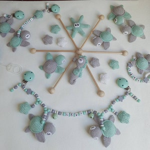 Può includere: Un mobile per neonati con una combinazione di colori verde e grigio. Il mobile presenta tartarughe marine, stelle marine e perline all'uncinetto. Il mobile è attaccato a una struttura in legno con una catena di perline grigie e bianche. La catena ha una perlina di legno con il nome "Levi" su di essa.