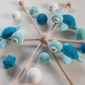 Könnte beinhalten: Ein Holz-Babymobil mit gehäkelten Meeresschildkröten- und Sternen-Anhängern. Das Mobile verfügt über drei blaue und grüne Schildkröten, zwei weiße Kugeln, zwei blaue Sterne und vier blaue und grüne Kugeln.