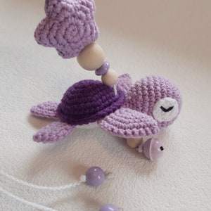 Puede incluir: Un juguete de tortuga de crochet morado con una estrella y cuentas de madera. La tortuga tiene un ojo blanco y una campana en la cola. El juguete est&aacute; unido a una cuerda blanca.