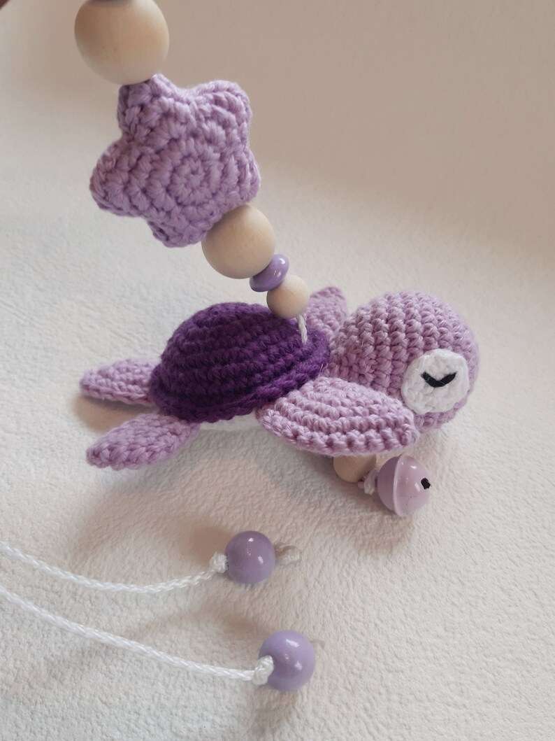 Puede incluir: Un juguete de tortuga de crochet morado con una estrella y cuentas de madera. La tortuga tiene un ojo blanco y una campana en la cola. El juguete est&aacute; unido a una cuerda blanca.