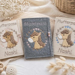 Op de afbeelding: Drie rechthoekige vilten hoezen voor medische boekjes. Twee zijn crèmekleurig met de tekst Mutterpass en Nashi, en één is donkergrijs met de tekst Mamipass. Elke hoes heeft een geborduurd bruin paard binnen een bloemenkrans met hartjes.