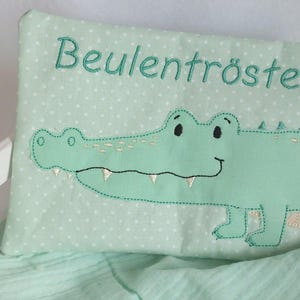 Könnte beinhalten: Ein hellgrünes Kissen mit weißen Punkten und besticktem Text, der "Beulentröster" lautet. Das Kissen zeigt ein besticktes grünes Krokodil mit weißen Zähnen und Augen.