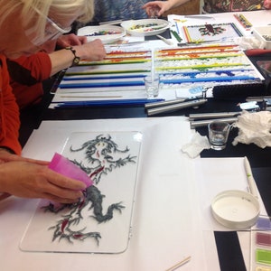 May include: A close-up of a glass panel with a black and white illustration of a dragon. The dragon is surrounded by red, white, and blue glass pieces. The panel is on a white surface with a pink tool and a wooden stick.