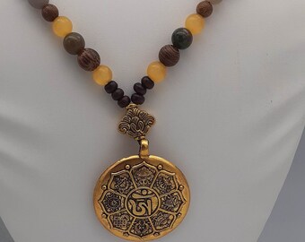 Könnte beinhalten: Halskette mit goldfarbenem Anhänger und Perlenkette. Die Perlen sind in Gelb-, Braun- und Grautönen gehalten. Der runde Anhänger hat detaillierte Muster und ein zentrales Symbol.