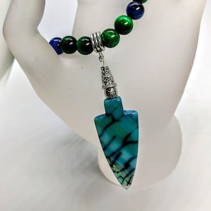 Puede incluir: Un collar de cuentas verdes, azules y negras con un colgante de plata con forma de punta de flecha. La punta de flecha está hecha de ágata azul y verde.