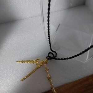 May include: A gold-colored necklace with a pendant featuring two stylized swords. The necklace is made of a black braided cord.