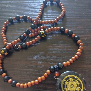 Puede incluir: Un collar de cuentas de madera marrón y negro con un gran colgante negro con un diseño geométrico dorado. El colgante es un disco redondo y plano con un patrón geométrico dorado.