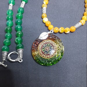 May include: A green and yellow beaded necklace with a round, multi-colored glass pendant featuring a flower and text. The pendant is attached to the necklace with a silver chain.