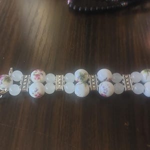 May include: A double-strand bracelet featuring white and light blue beads. The bracelet has floral-patterned ceramic beads and silver-toned metal accents. The bracelet is approximately 7 inches long.