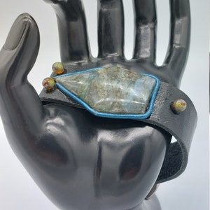 May include: A black leather bracelet with a large, gray and blue gemstone set in a silver-toned bezel. The bracelet is accented with small, colorful glass beads.