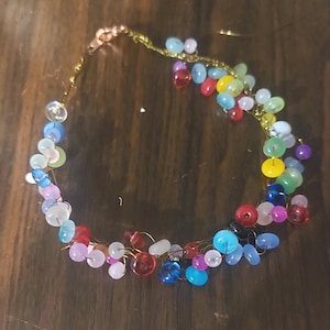 May include: A colorful beaded necklace with a gold-colored chain. The necklace features a variety of round and oval beads in shades of red, blue, green, yellow, and pink. The beads are strung together to create a unique and eye-catching accessory.
