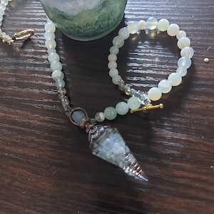 May include: A light blue and green beaded necklace with a crystal point pendant. The necklace is made with natural stones and has a gold clasp. The pendant is a clear crystal with a silver wire wrap.