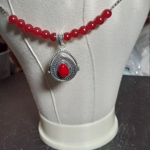 May include: A silver necklace with a red gemstone pendant. The pendant is a locket with a detailed design. The necklace is made of red beads and a silver chain.